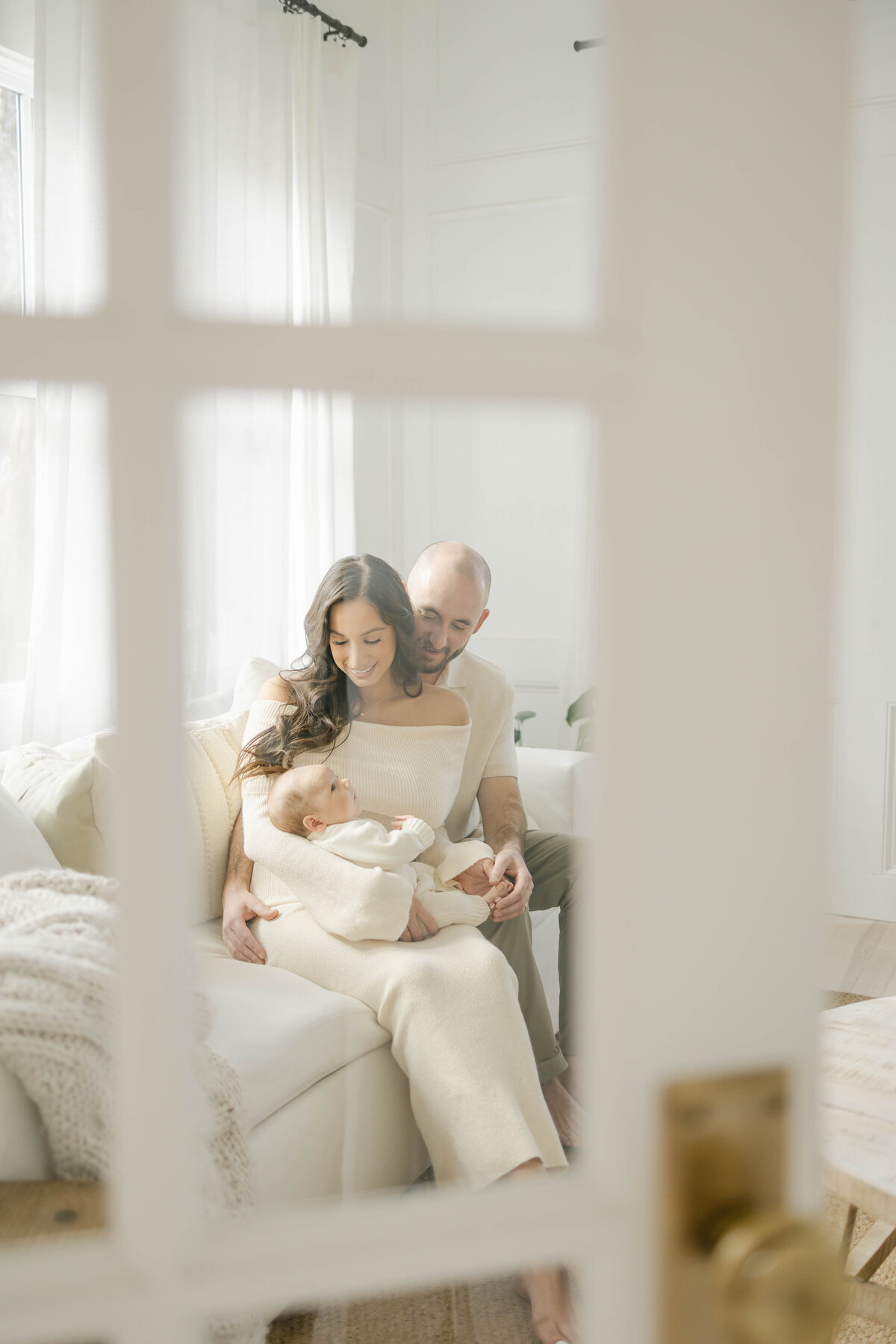 a mom and dad looking at their baby while sitting in a white living room in dallas texas, view through a window