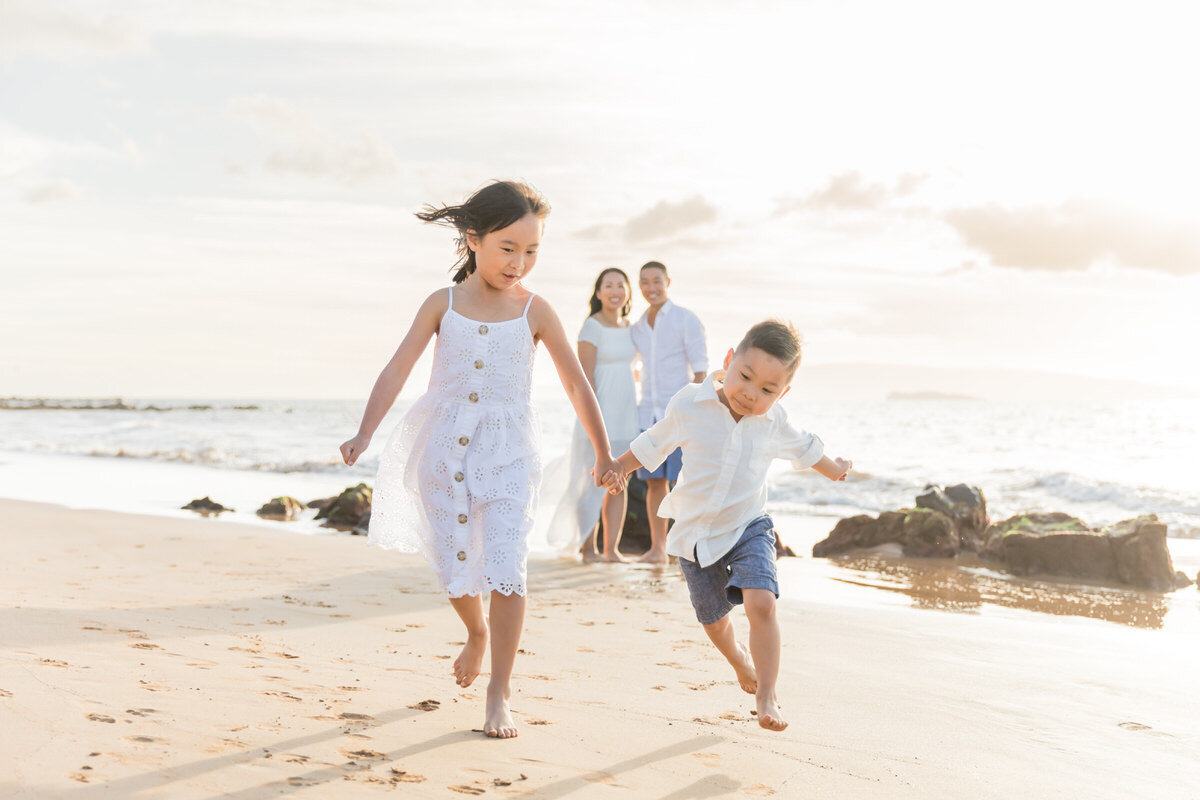 Little boy and girl running hand-in-hand down the beach with mom and dad looking on. Everyone has smiling faces.