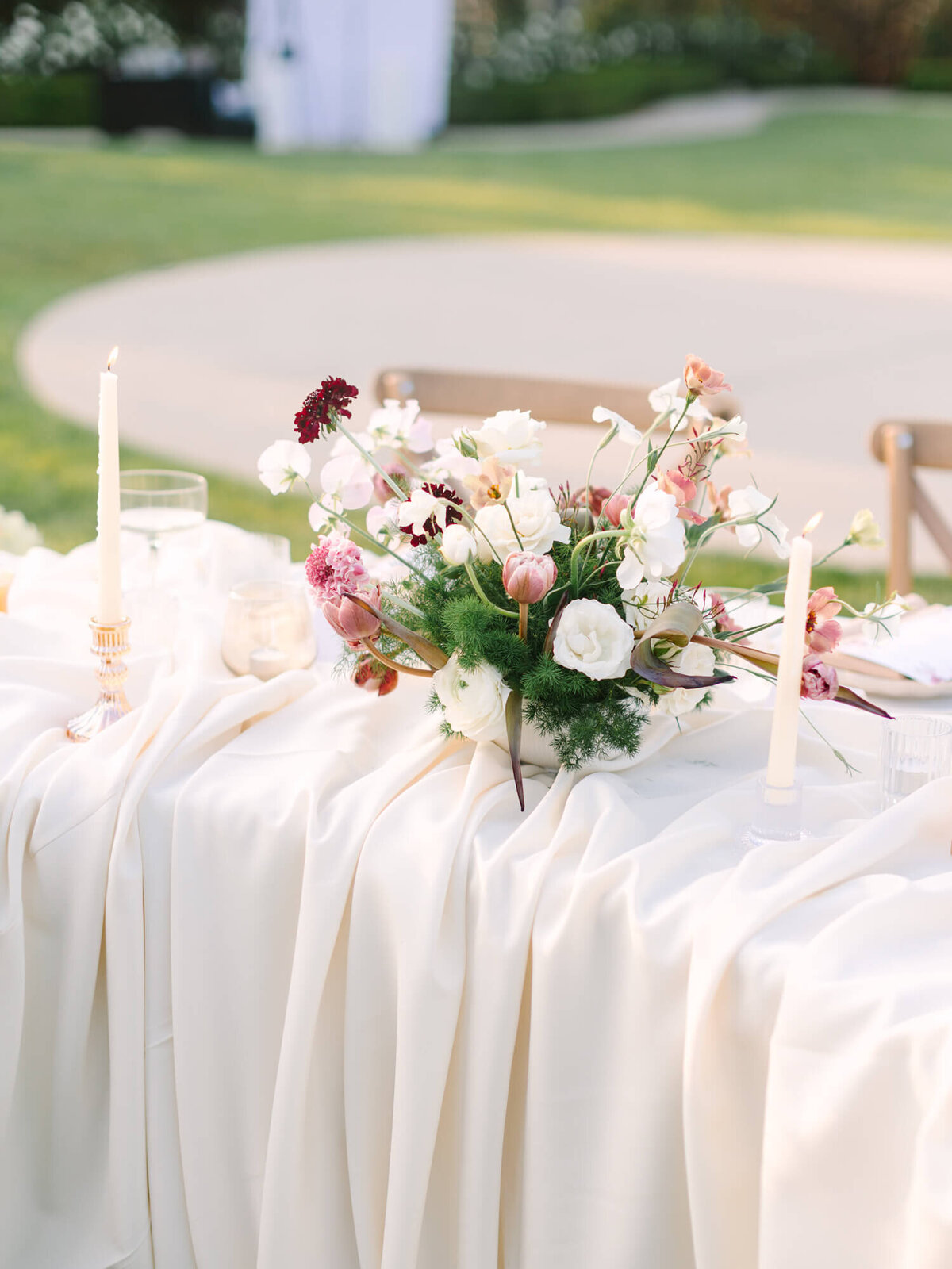 Elegant outdoor wedding table with white cloth, adorned with a lush floral centerpiece of pink and white flowers and tall, lit candles.