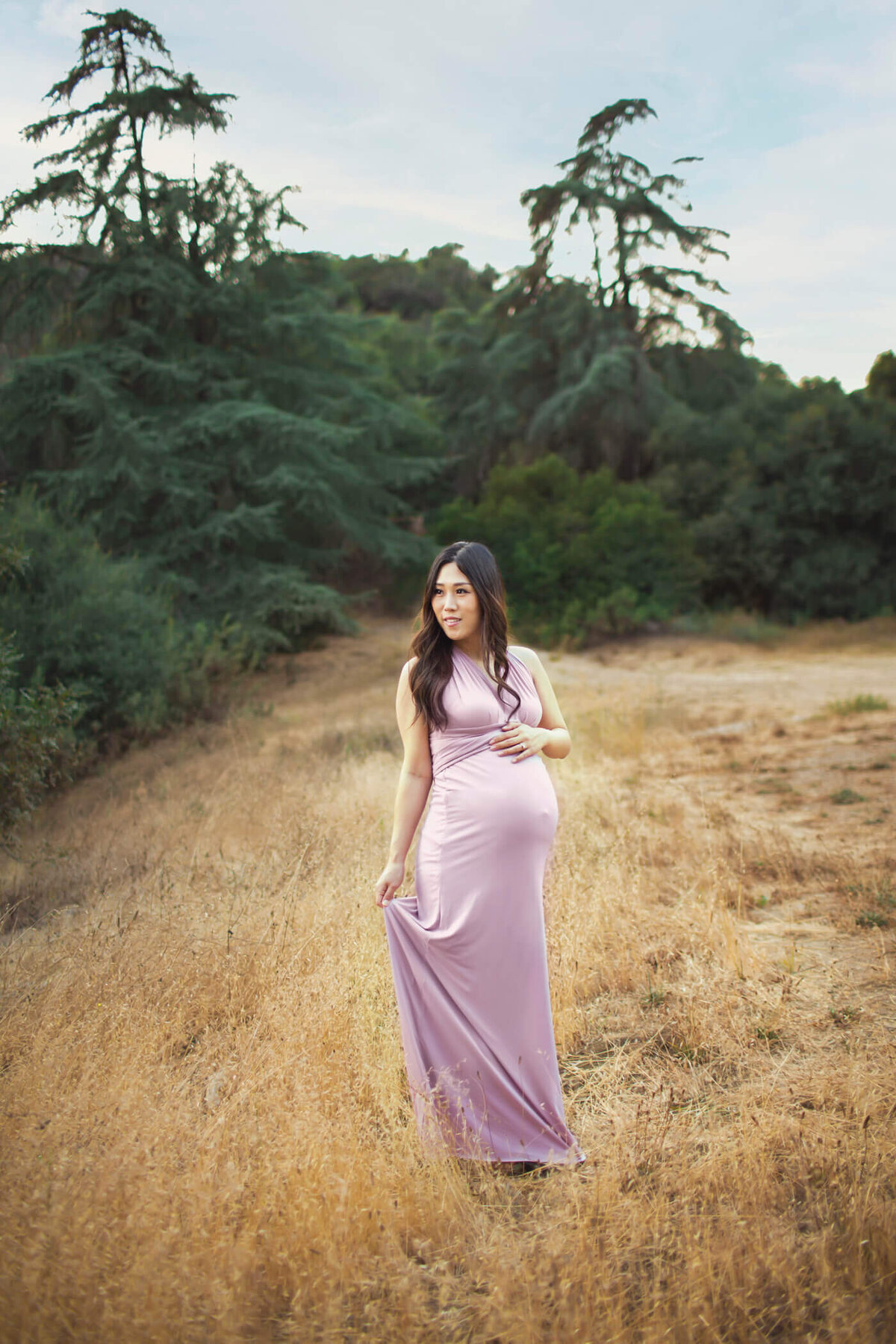 A pregnant woman stands in a golden field, surrounded by lush greenery and tall trees. She is wearing a form-fitting, sleeveless lavender dress that highlights her baby bump. With one hand resting on her belly and the other holding the hem of her dress, she gazes thoughtfully into the distance. The natural setting and soft lighting create a serene and peaceful atmosphere.