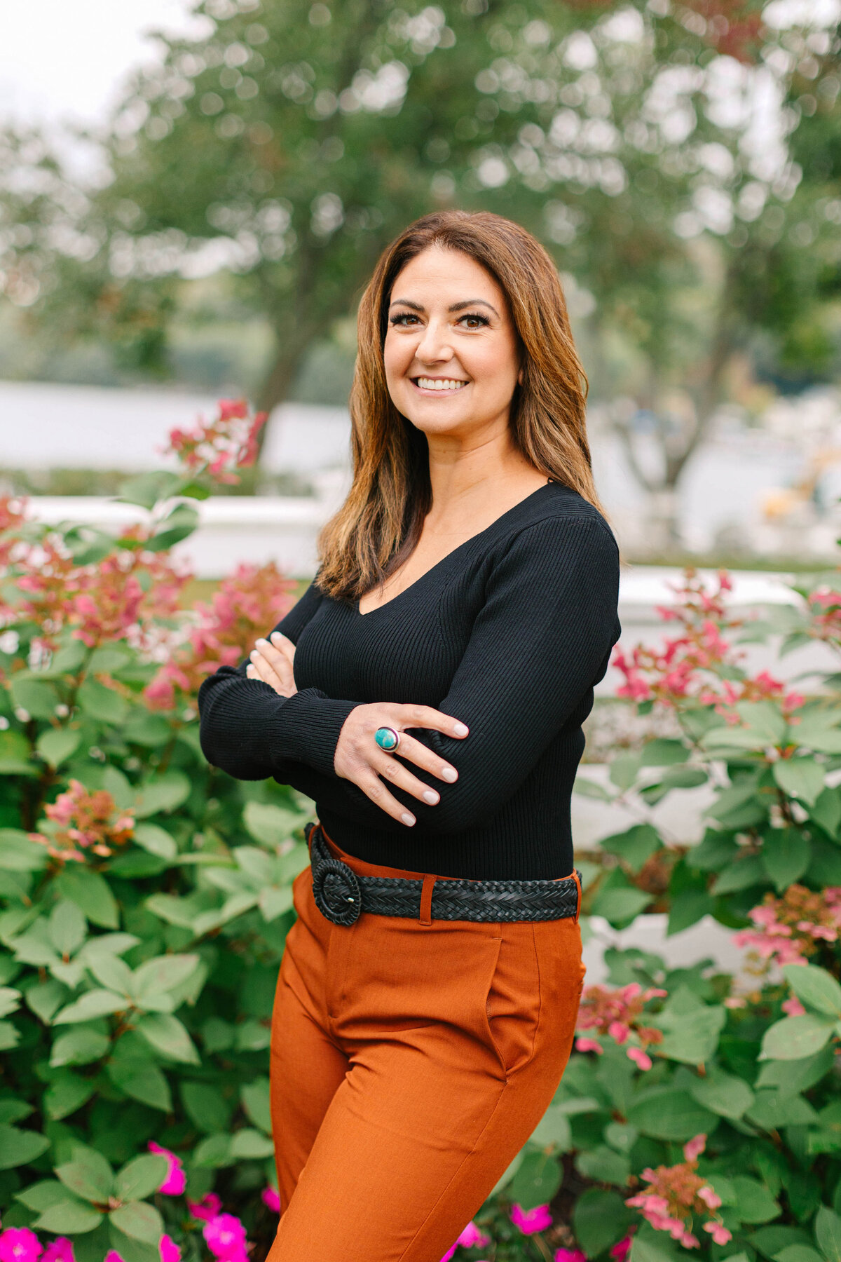 A confident woman with shoulder-length brown hair stands with her arms crossed in front of lush greenery and pink flowers. She wears a black ribbed top, rust-colored trousers, a woven belt, and a turquoise statement ring, with a lake and trees in the background.
