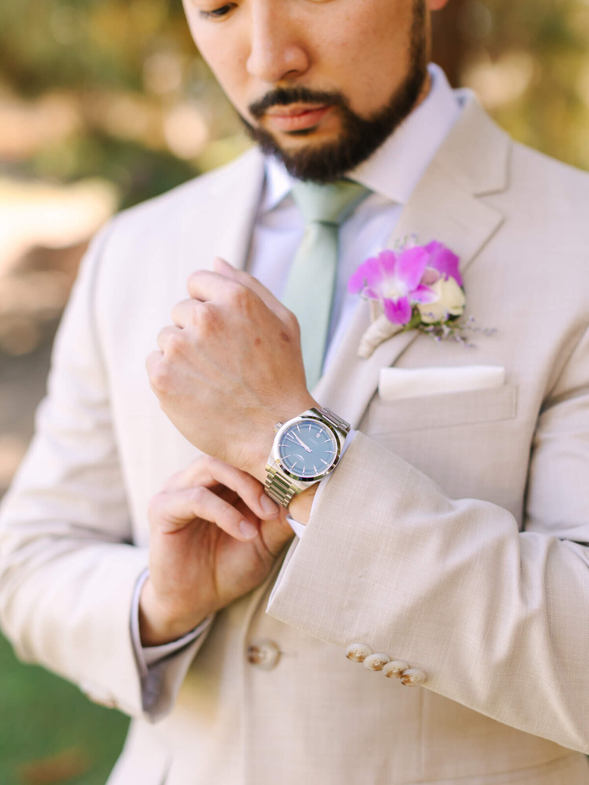 Groom in a beige suit with a green tie adjusts a silver watch. A pink flower boutonnière adorns his lapel. The scene feels elegant and formal.