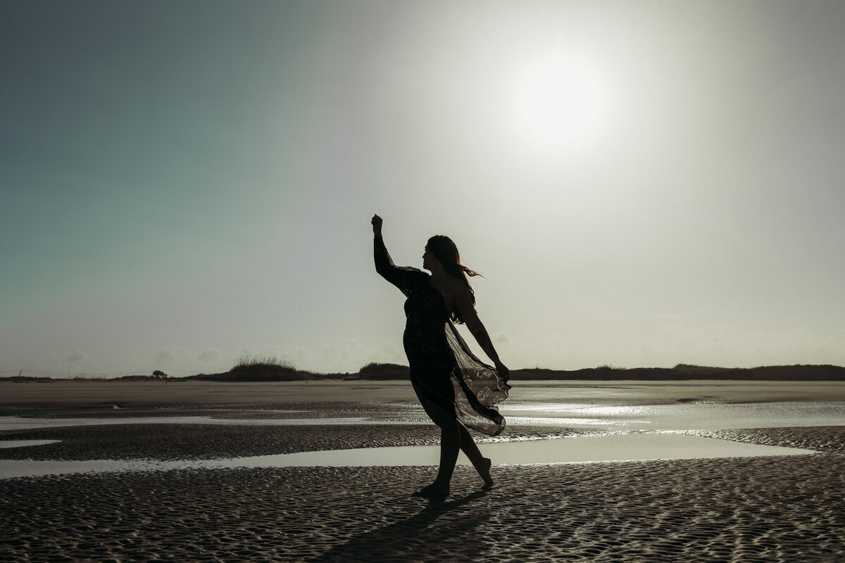Silhouetted woman walking barefoot on a beach with flowing sheer robe at sunset in a maternity-style beach portrait session.