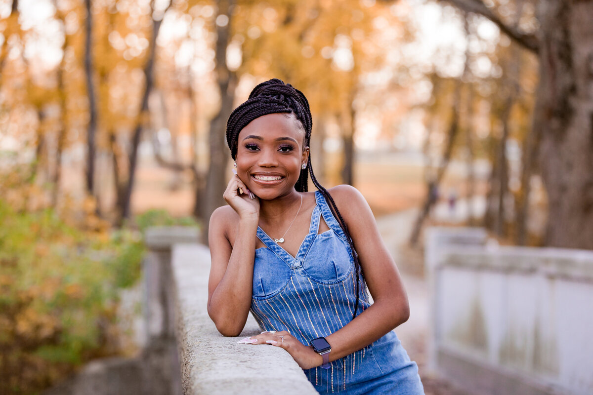 A young woman in braids and a blue jean dress leans on a wall sideways with her hand under her chin.