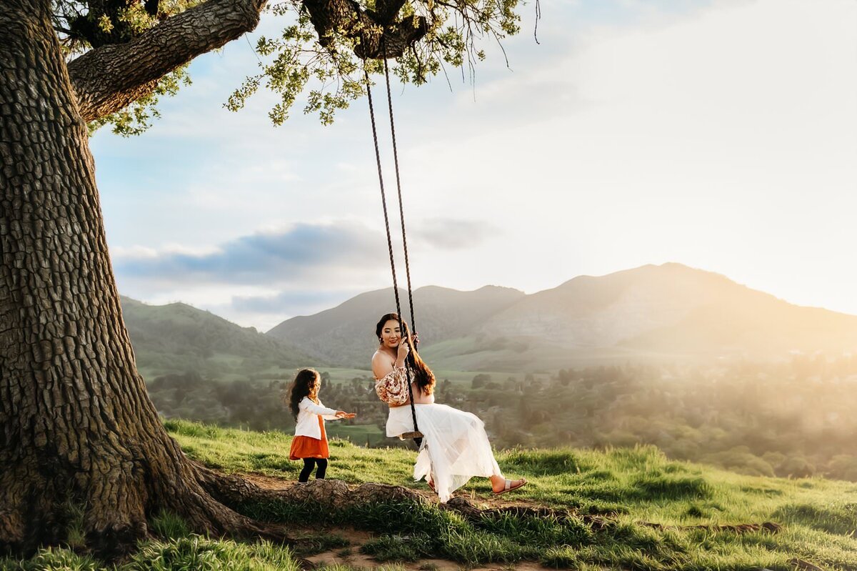 little girl pushed her mom on a tree swing. the mom is pregnant and wearing a 2 piece maternity gown