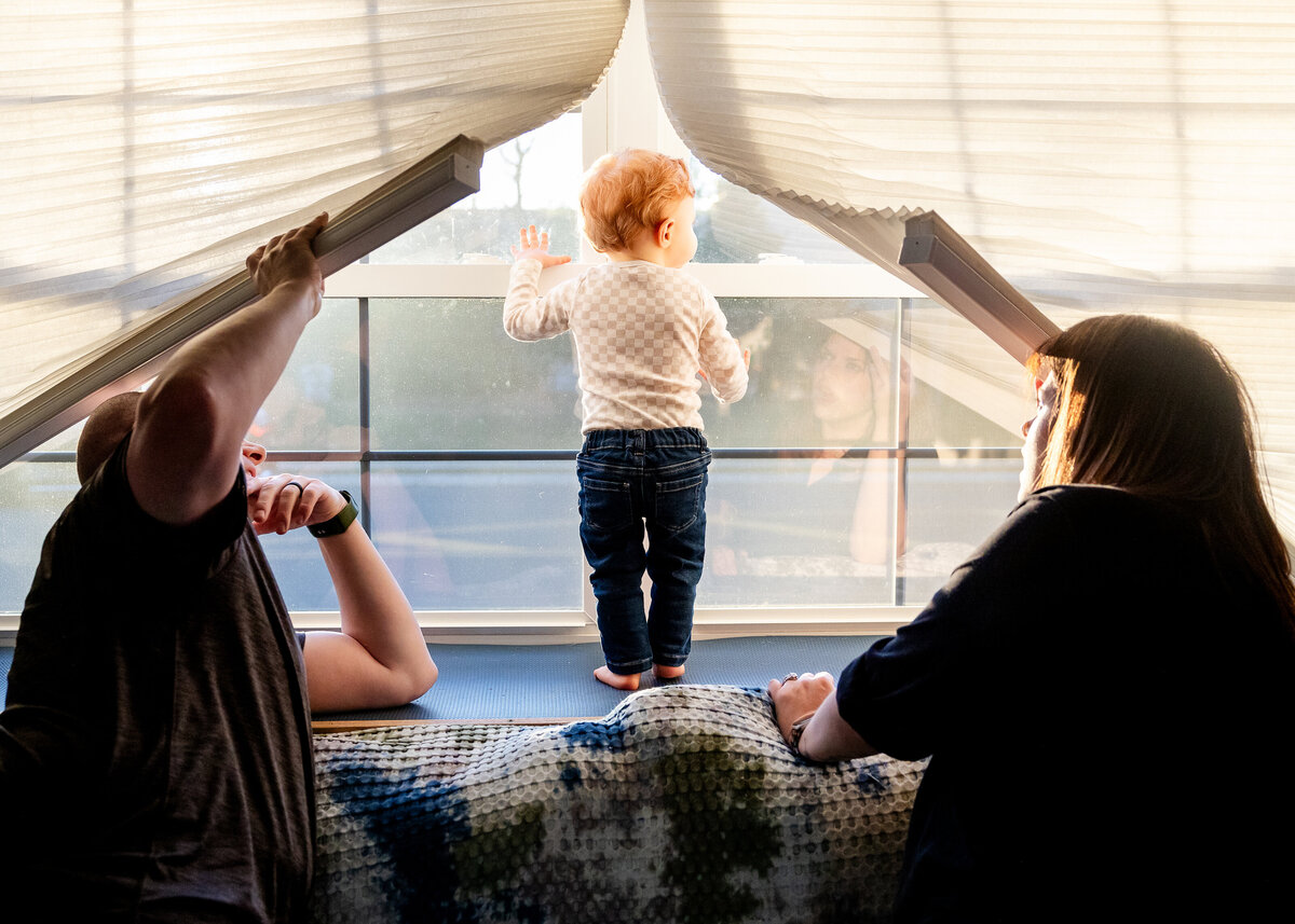 Toddler playing on windowsill while mom and dad watch and lift window shades