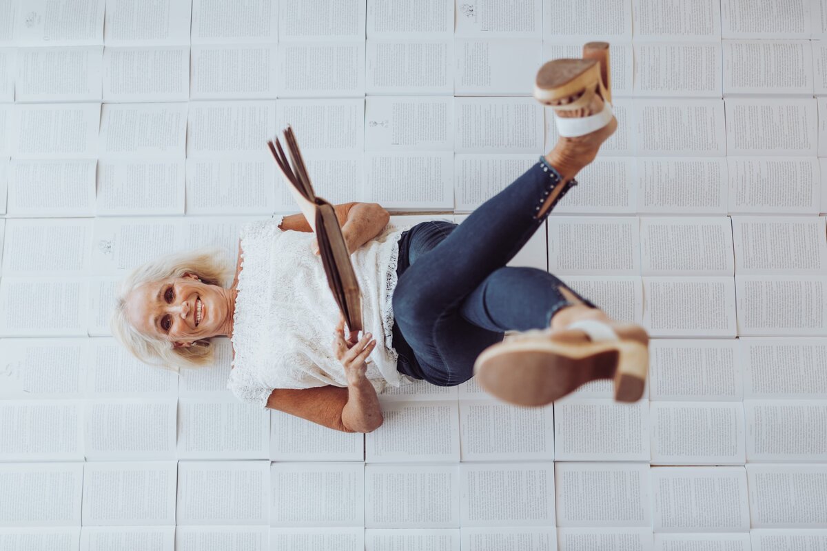 Woman wearing a white top, jeans and heels lays on a bunch of open books and smiles at the camera, holding a book in her hands.