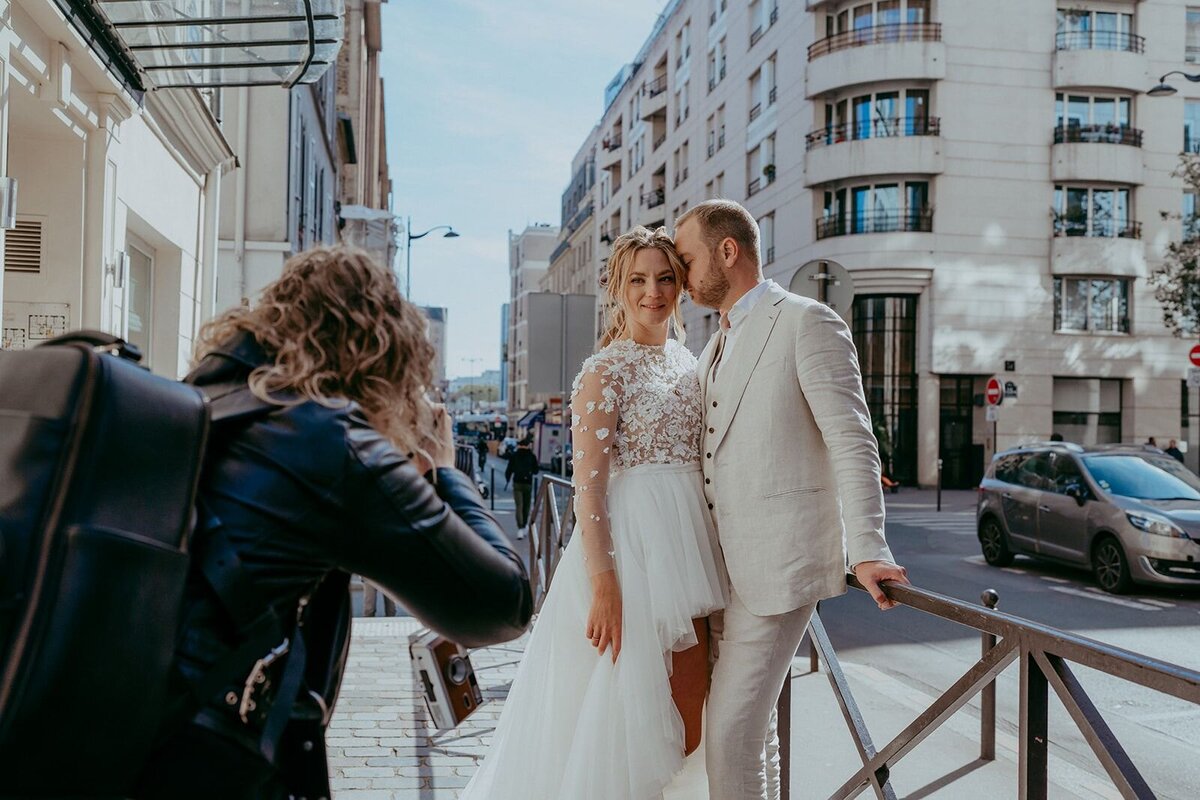 Fotograaf met zwart leren jasje en zwarte leren tas fotografeert bruidspaar in Parijs bruid jurk voor kort en achter lang bruidegom lichtbeige pak aan