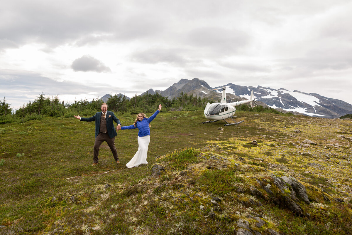 A bride and groom walk down a green hill with their hands in the air celebrating.