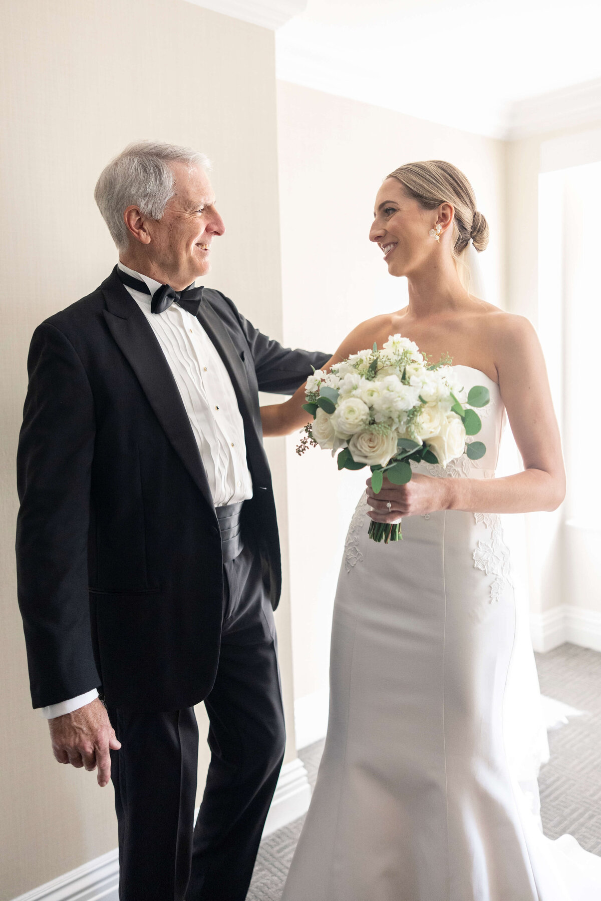 A bride in a strapless wedding dress holds a bouquet of white flowers, smiling warmly at her dad in a black tuxedo. They stand indoors with bright natural light coming through a nearby window.