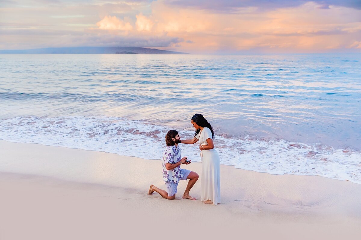 Man in white and blue shirt gets down on one knee on the beach in Maui to propose to the love of his life