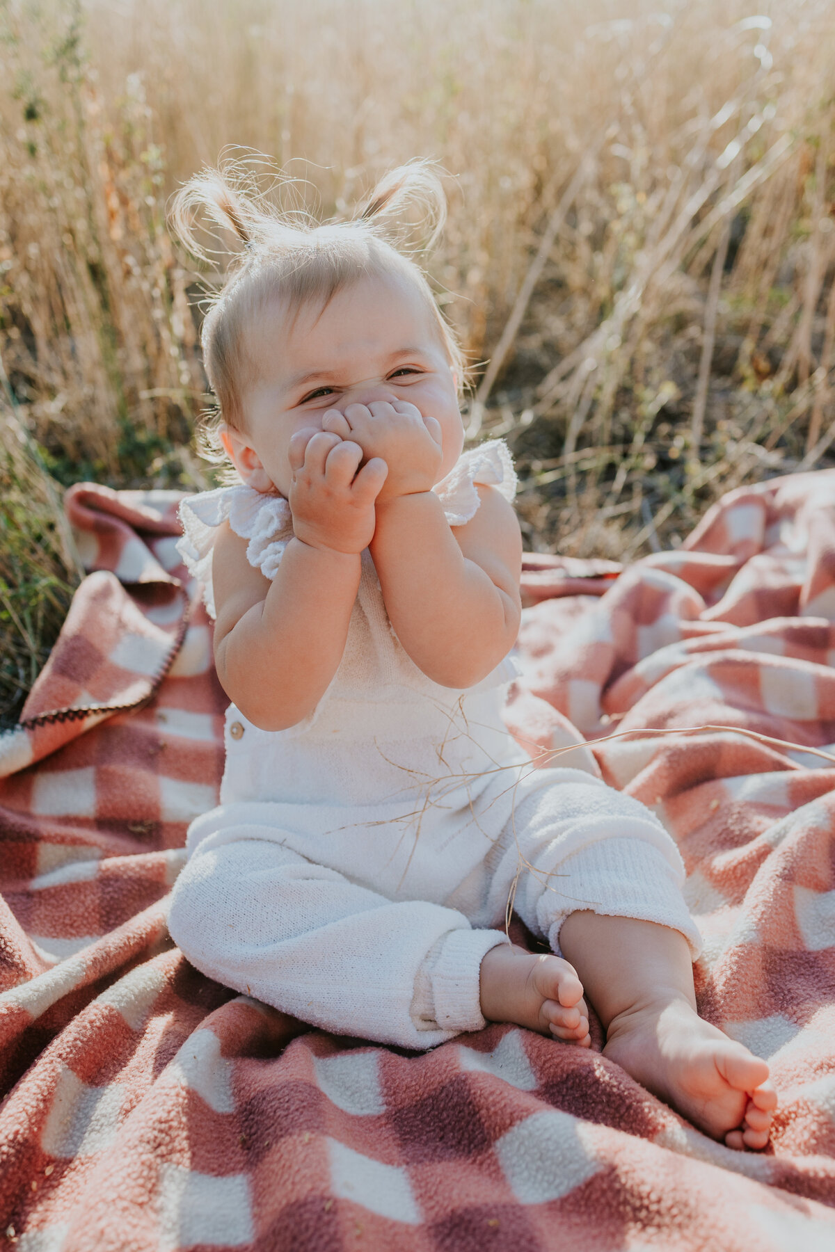 One year old girl covers her mouth and giggles at camera while sitting on red checked blanket in golden field.