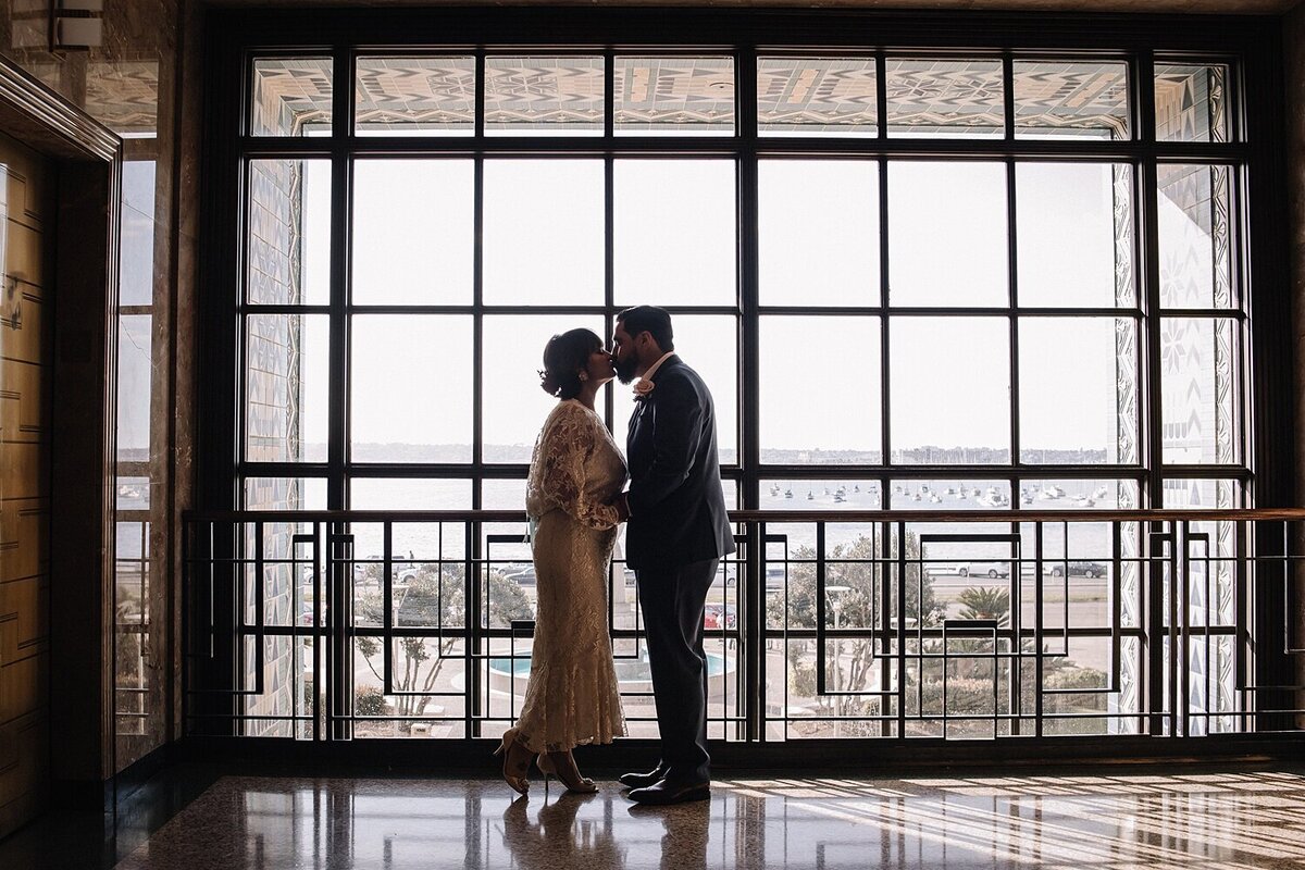 photo of a couple kissing. they are standing on front of a floor to ceiling window in the tower of the san diego county administration building