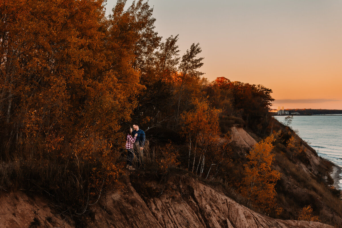 man and woman snuggle on the shores of lake michicagan at Lions Den Gorge while fall foliage surrounds them