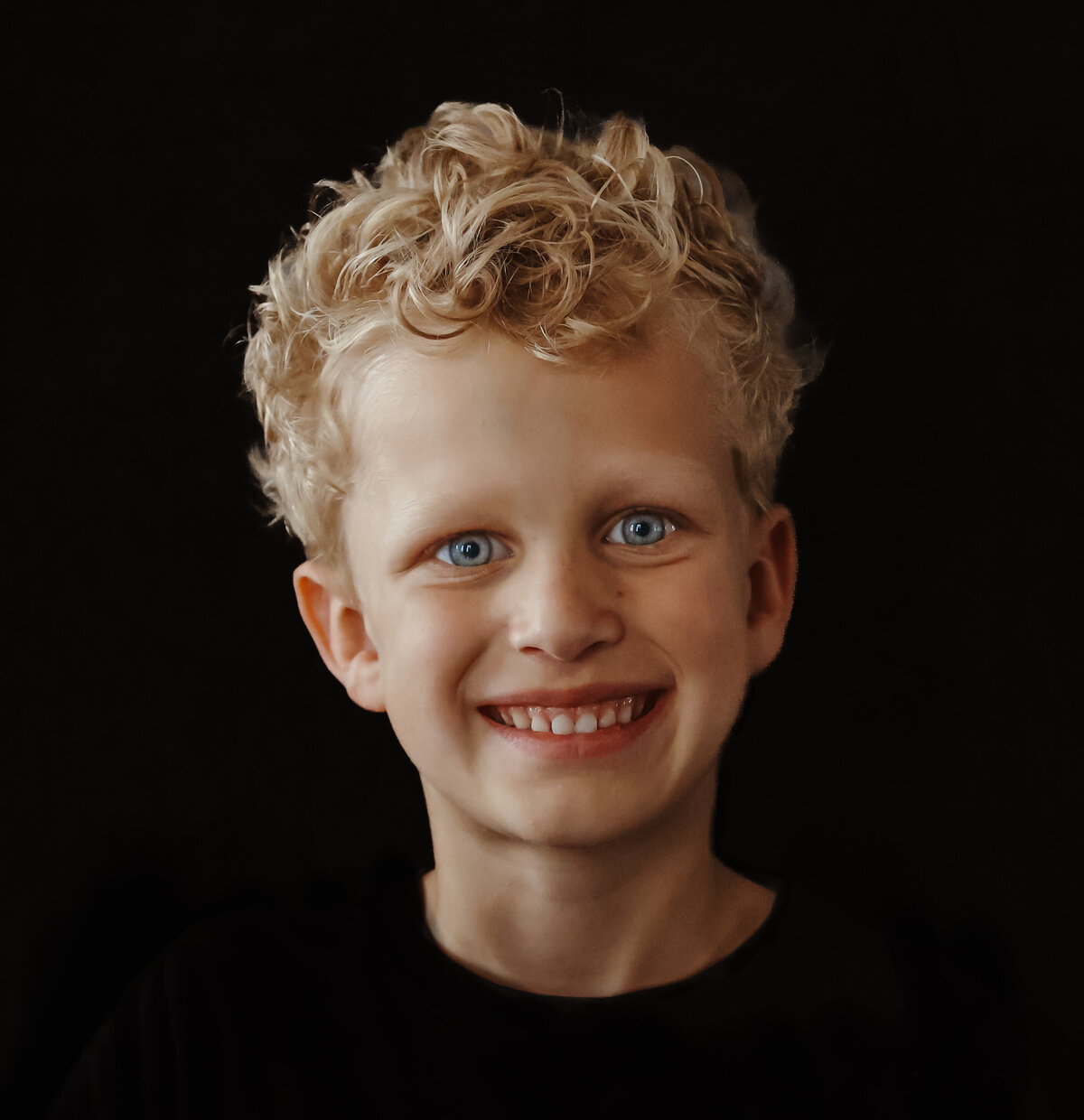 een portret van een vrolijke jongen met krullen tegen een zwarte achtergrond
