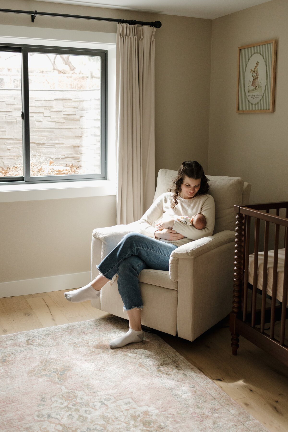 New Longmont Colorado mom holding newborn in rocking chair and looking down at baby with legs crossed 