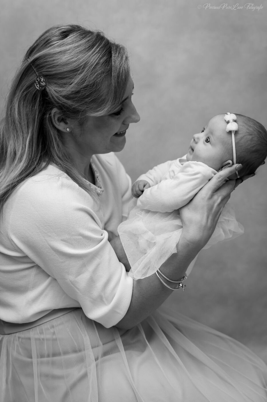 A black and white, medium shot of a mother holding her newborn baby. The mother, smiling down at the baby, has her hair in a half-up style and is wearing a sweater with a beaded collar and a light-colored dress. She holds the baby, who is looking up at her with a curious expression.