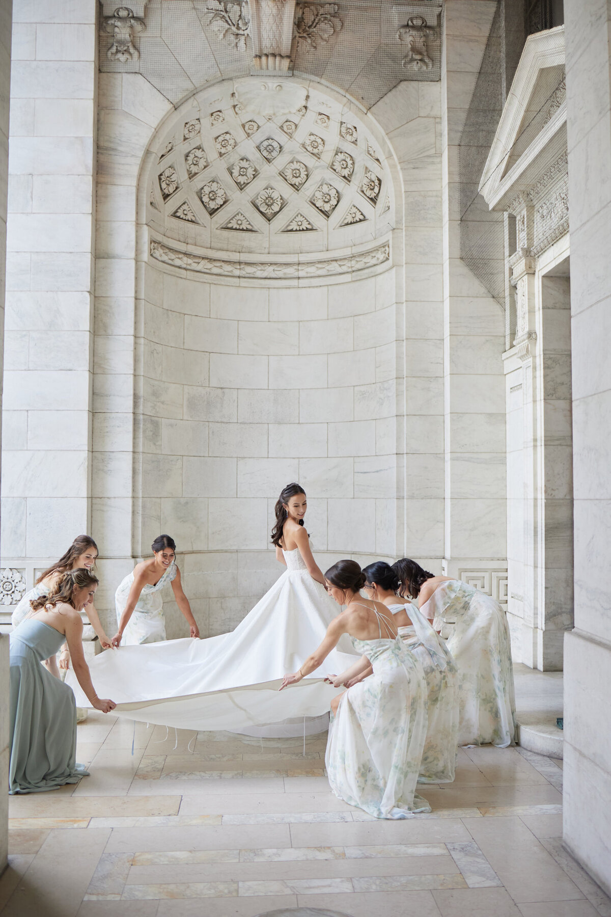 The photo was taken at The New York Public Library in New York City by photographer Shawn Connell with videography by Aaron Navak Films. The scene captures a bride in a white gown standing in an ornate, white marble hallway, with five bridesmaids delicately arranging her long train. The setting exudes elegance and grandeur with its detailed architecture and high ceilings.