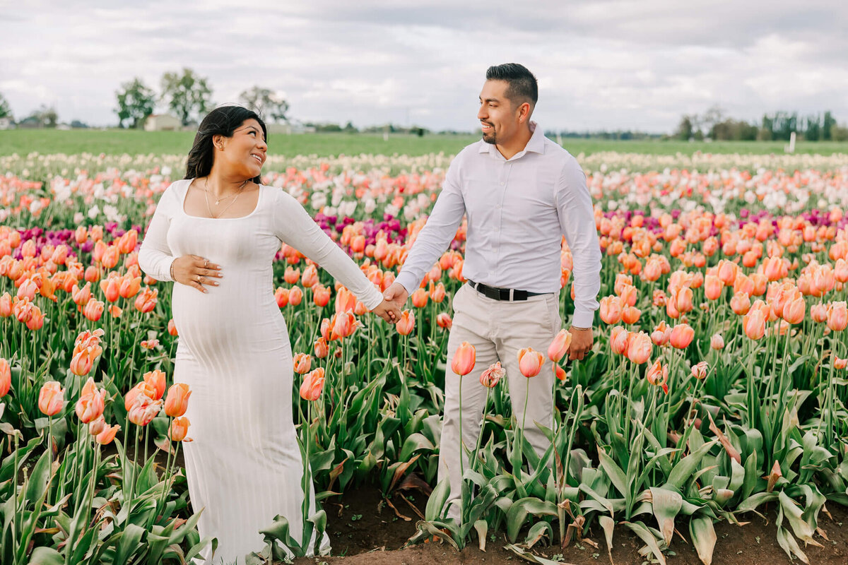 Portrait of a husband and wife walking in the tulip fields in woodburn oregon. She is wearing a white dress and has her hand on her belly and he is smiling at him behind her.