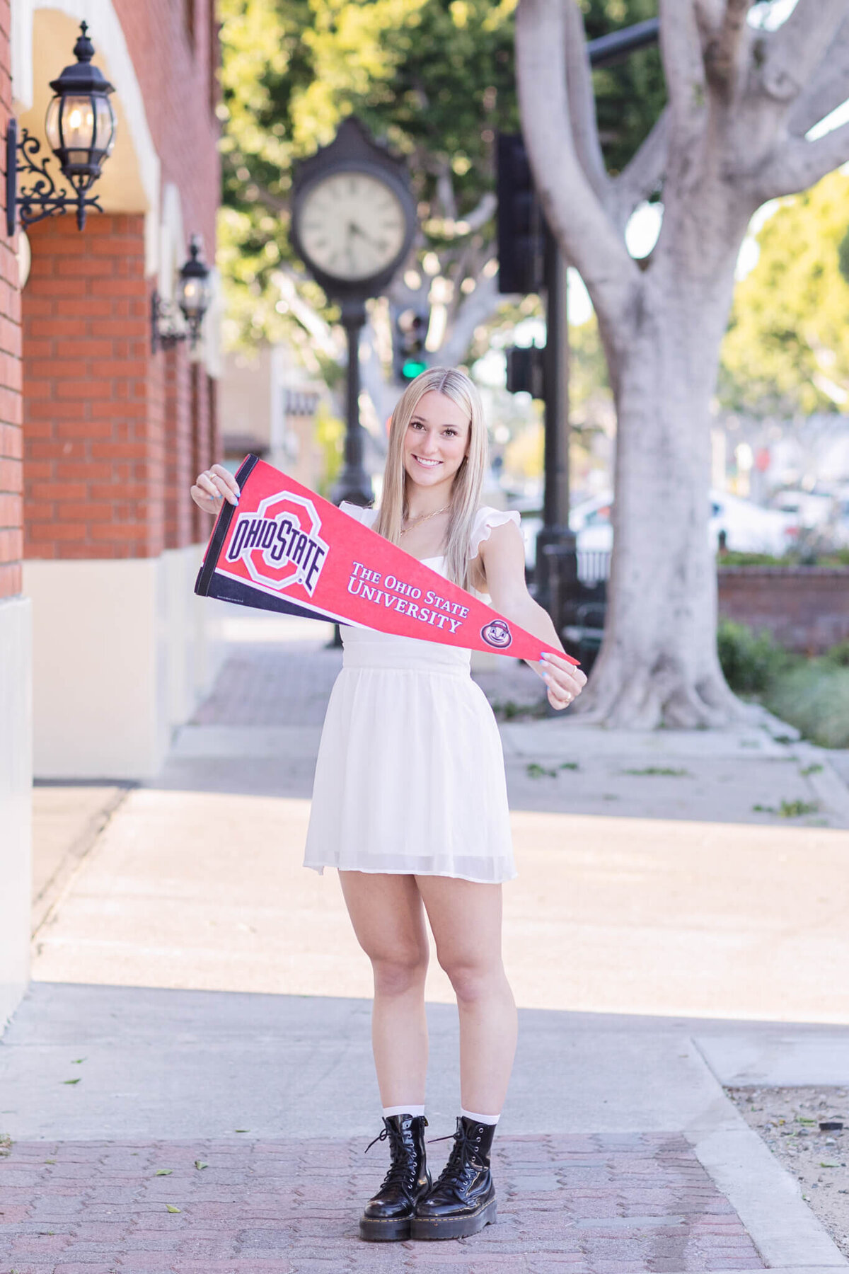 A blonde woman in a white dress and black boots stands on a sidewalk holding an Ohio State University banner. There are trees and a brick building in the background.