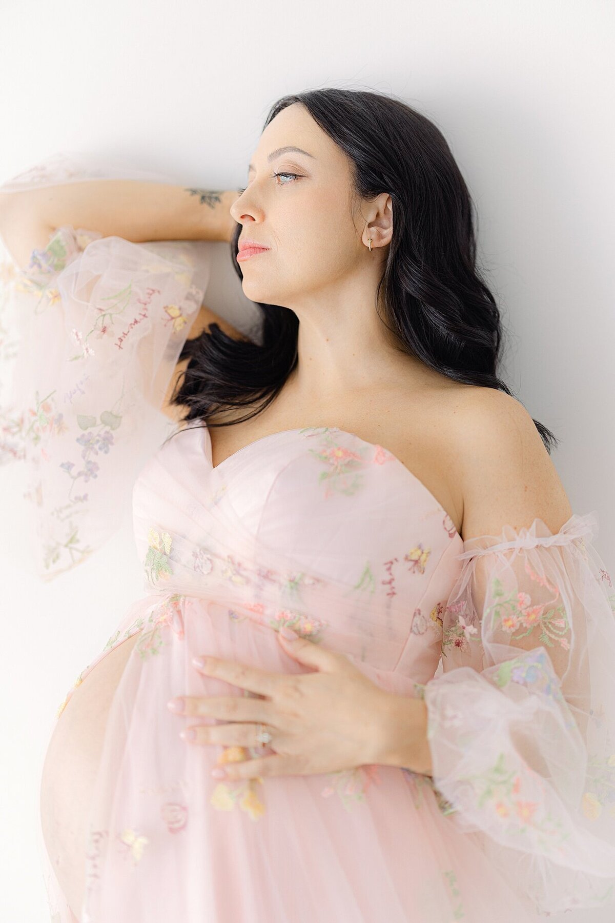 A pregnant woman with dark brown hair and a pink dress relaxes against a wall while holding her baby belly for her Indianapolis maternity photos