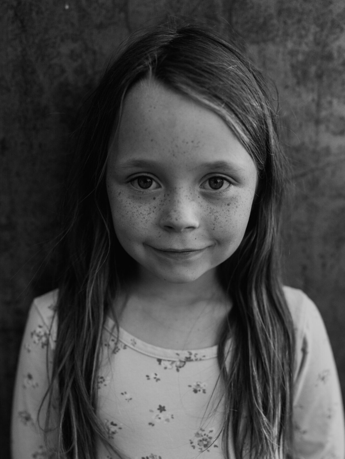 A close-up black-and-white portrait of an adorable girl, standing with her back to a metal wall on her family's farm.