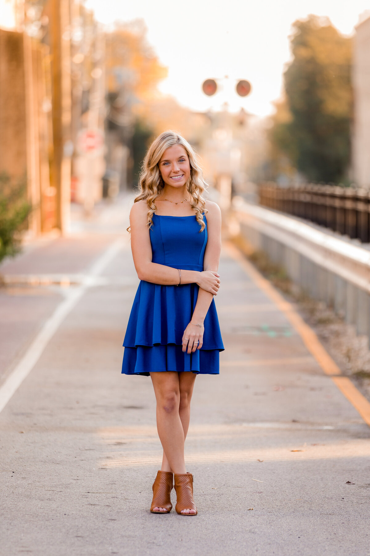 Kailey stands in the alley in a blue dress with her ankles crossed.