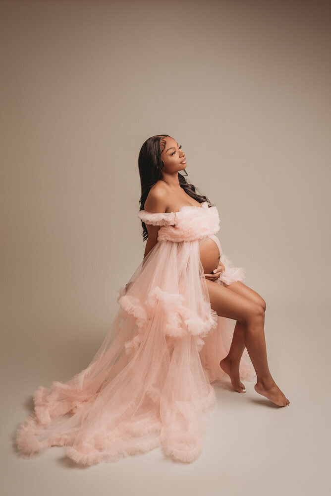 Atlanta, Georgia maternity photographer photographs 36 week pregnant woman at atlanta ga maternity portrait studio posing on stool in pink ruffle tulle dress holding baby bump on light grey backdrop