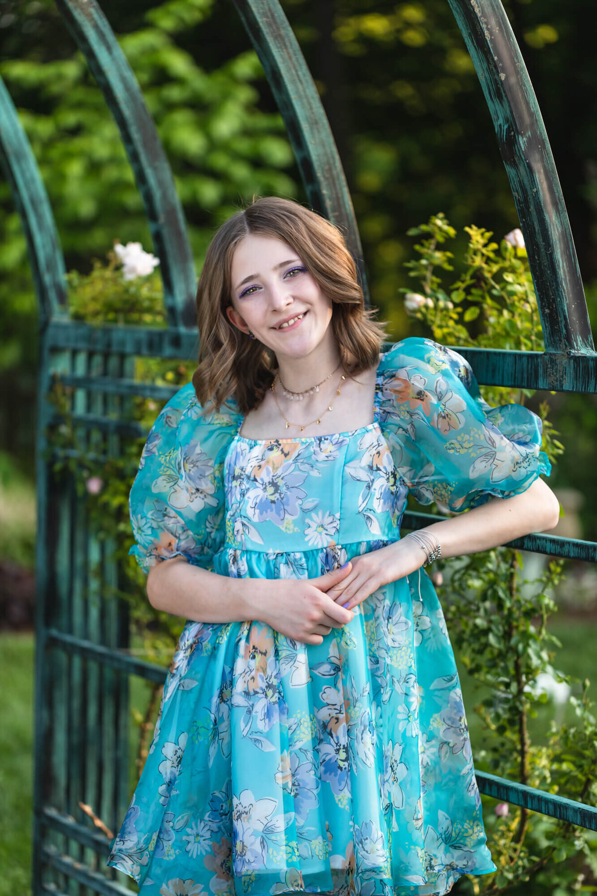 Pretty teenage girl in a blue floral dress with puff sleeves leaning on a green garden trellis surrounded by roses. Captured by Springfield, MO teen photographer Dynae Levingston.