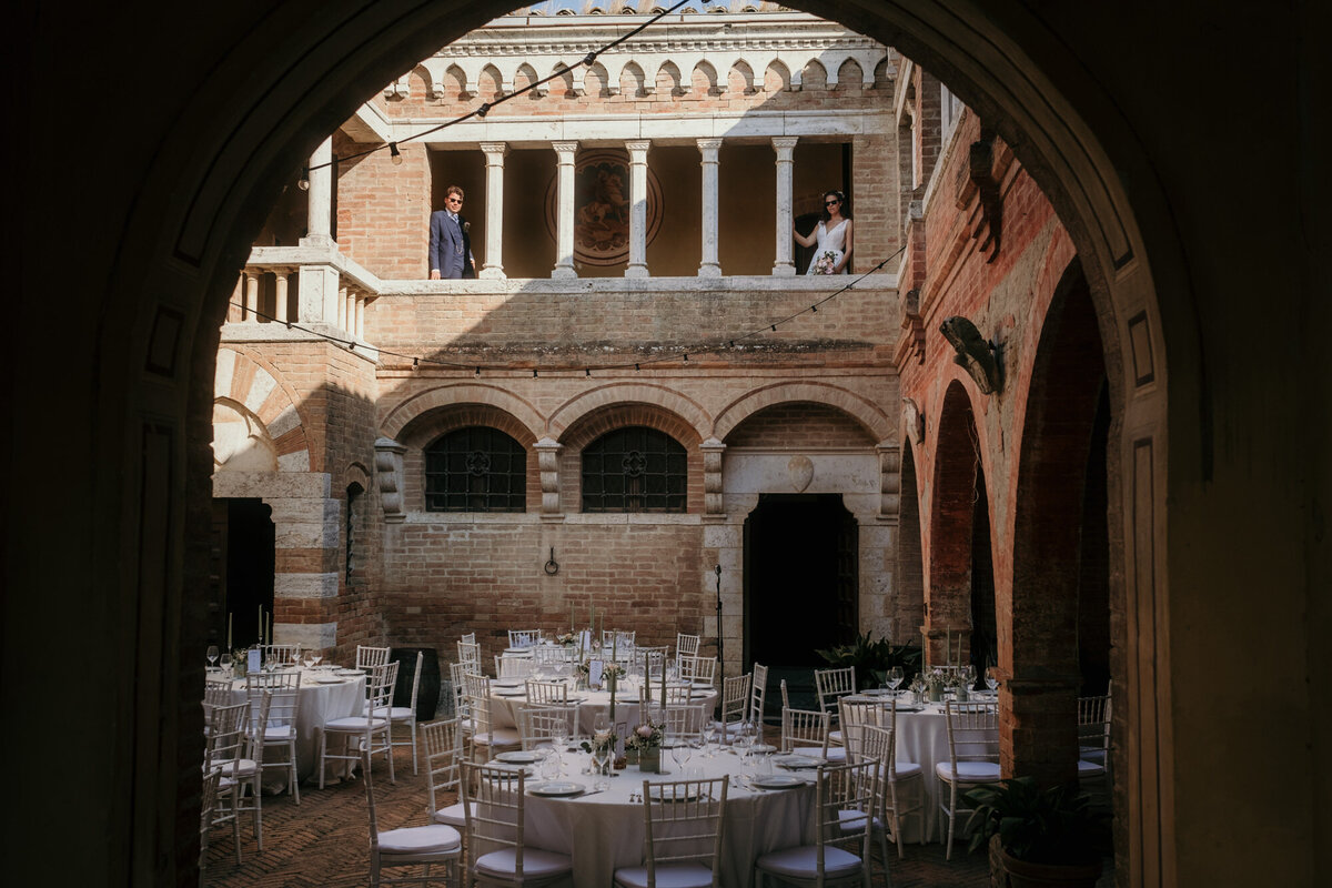During the wedding session bride and groom stand on a balkony looking down on their wedding venue with all prepaired tables.