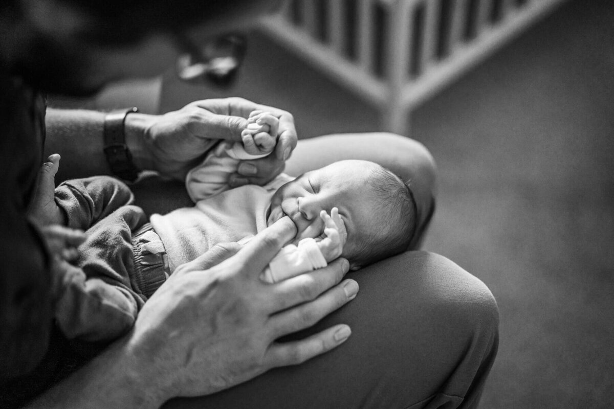 A black and white image of a newborn lying on his father's lap, being comforted by a finger substituting for a pacifier.