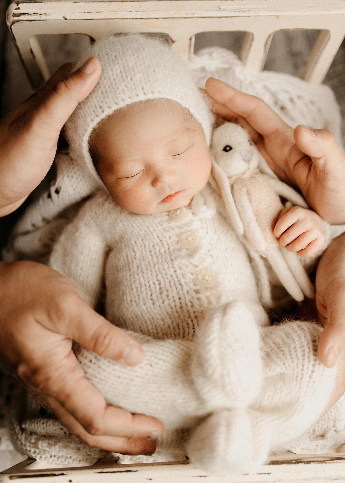 Newborn baby peacefully sleeping in a cream knit outfit and bonnet, holding a small stuffed bunny, while surrounded by the gentle hands of mom and dad.