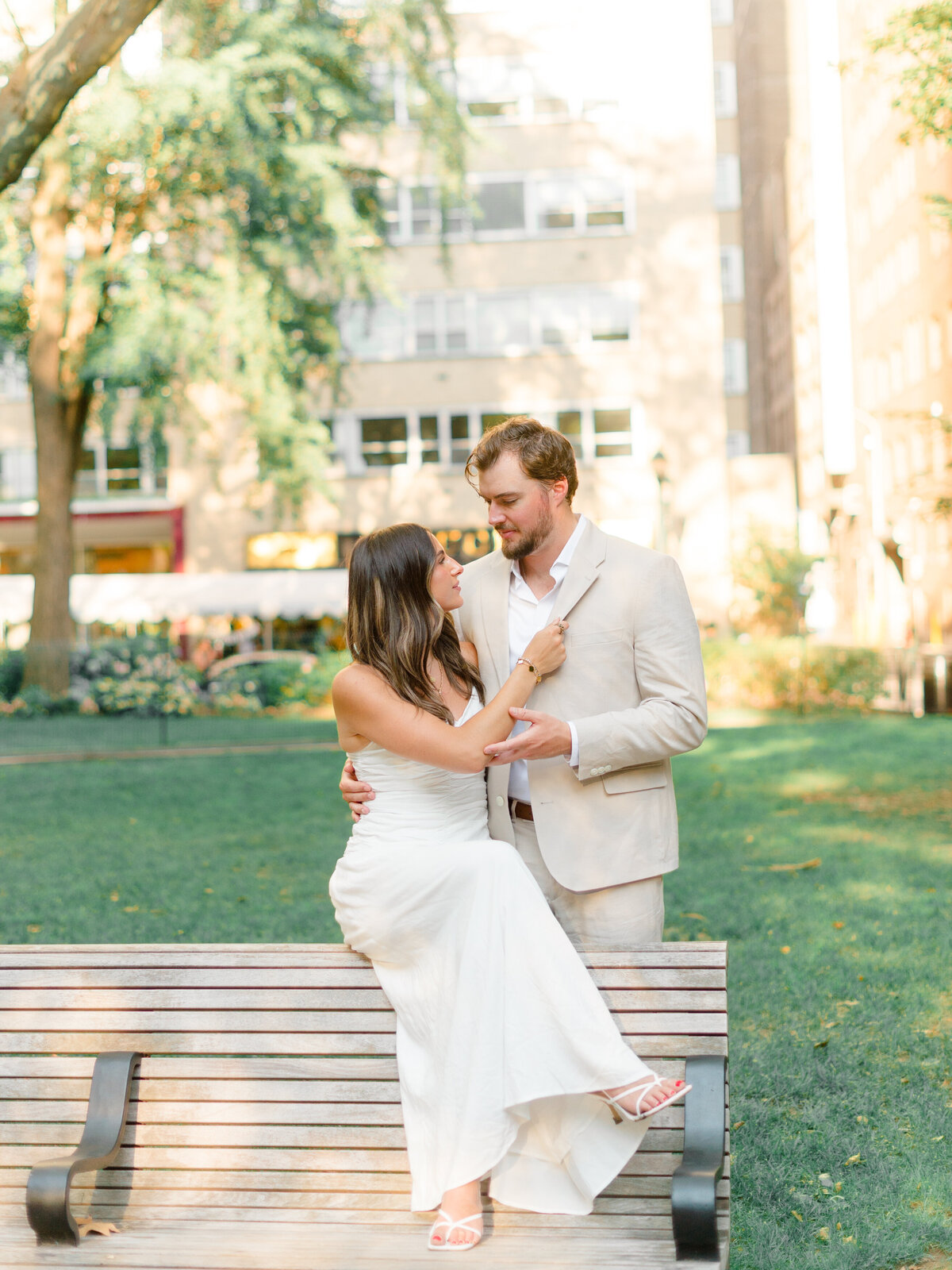A couple shares a quiet, intimate moment on a park bench in Rittenhouse Square. The woman sits elegantly in a white dress, gently resting her hand on her partner's chest, while he stands close, their eyes locked. Captured in warm, golden light by Elizabeth Kane Photography, a Luxury Philadelphia Wedding Photographer.