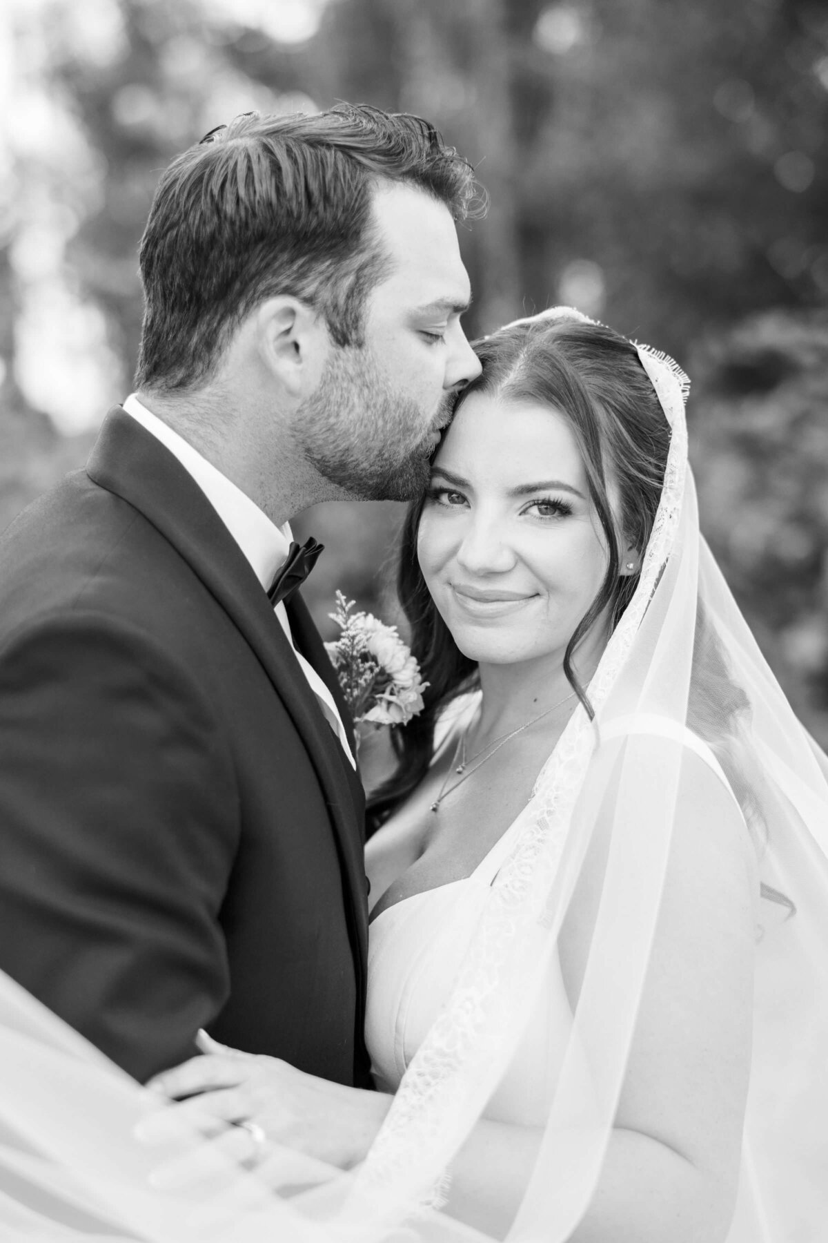 A bride smiles at the camera while the groom kisses her forehead at their outdoor luxury wedding in Oregon.
