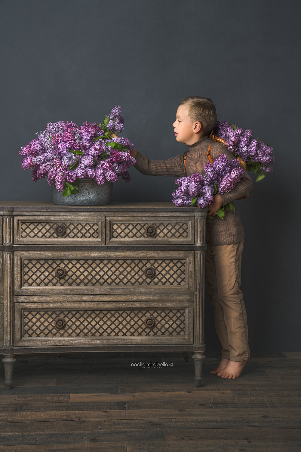 Boy standing on tip toes on edge of wooden dresser arranging fresh lilc blooms in a metal tub.