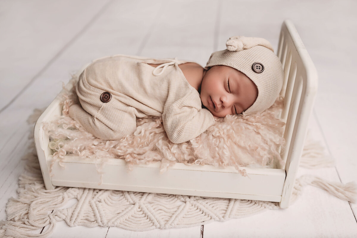 Newborn baby boy in cream sleeper outfit and cap laying on tiny white baby bed