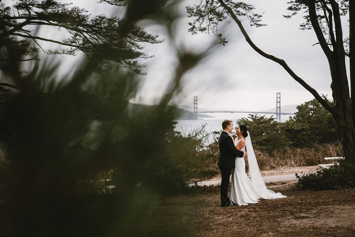 Bride and groom embrace outdoors with the Golden Gate Bridge in the background, framed by trees and natural scenery.