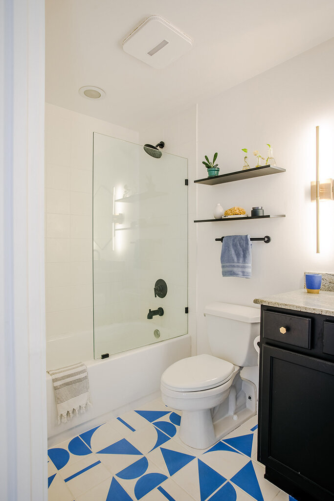 A modern bathroom with white walls and a striking white and blue geometric tiled floor. A white toilet sits next to a glass-enclosed bathtub with a black showerhead and fixtures. Two black floating shelves above the toilet hold small plants and toiletries. A light blue towel hangs from a black towel bar. A black vanity with a light countertop and gold hardware is visible on the right.