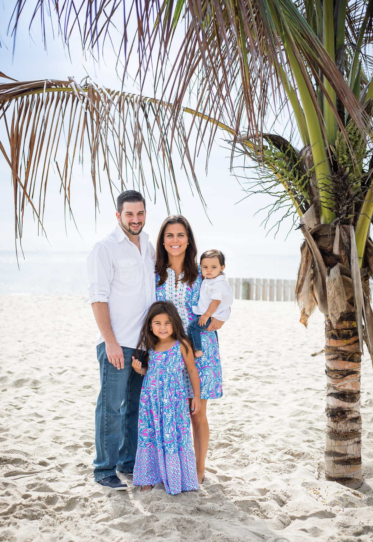 family of four standing under a palm tree on the beach in blue dresses