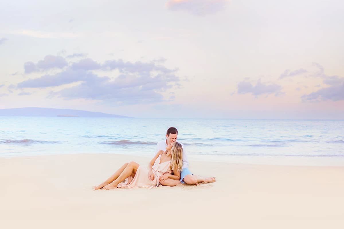 Couple sits on the sand at White Rock Beach on the island of Maui and kisses, woman is wearing a pink dress and sand is smooth