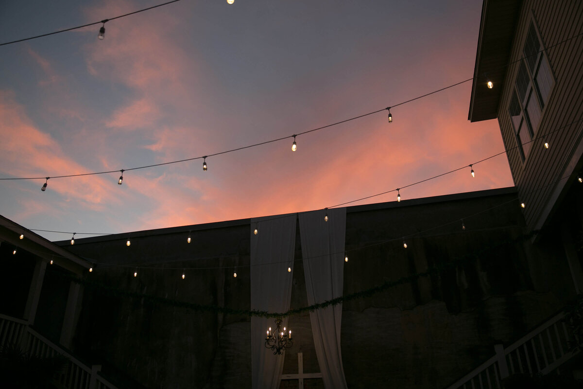 String lights hang above an outdoor space at sunset, with pink clouds in the sky. A white curtain and chandelier are set against a wall, partially framed by two buildings.