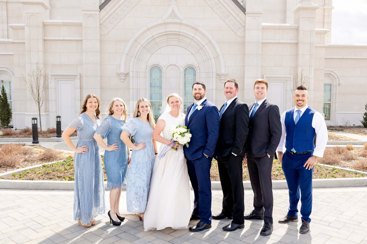 The bride and groom pose with their bridesmaids and groomsmen in coordinated blue outfits in front of the LDS temple.