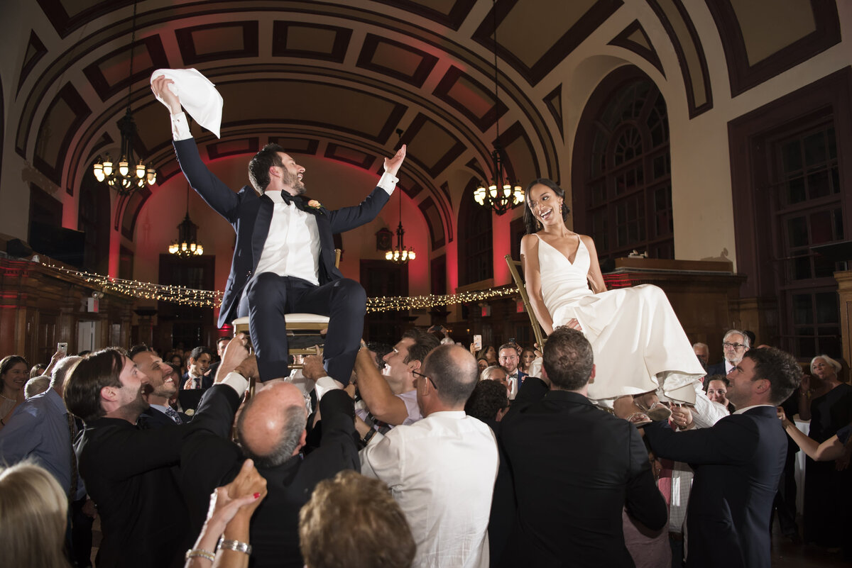 A newlywed couple being help up in chairs by their wedding guests