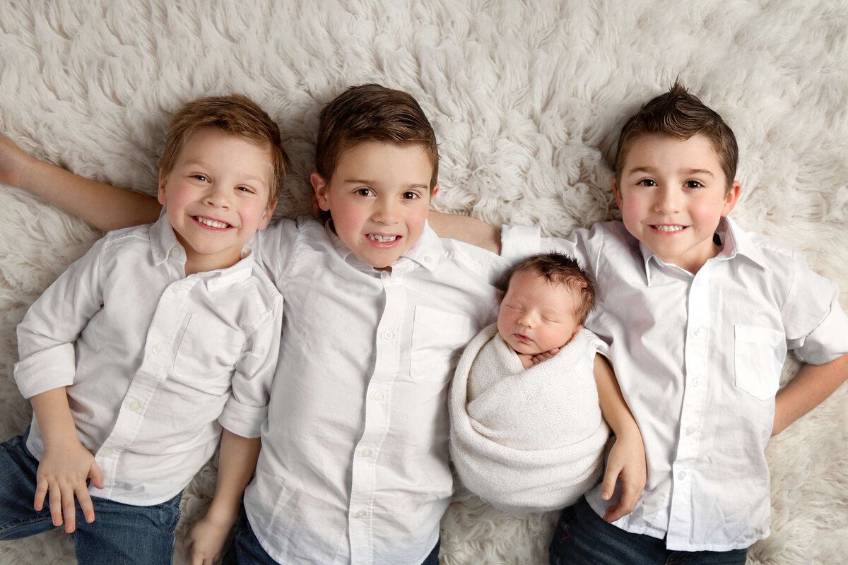 three big brothers in white button-up shirts with their newborn brother laying together on a white fur rug in a newborn session
