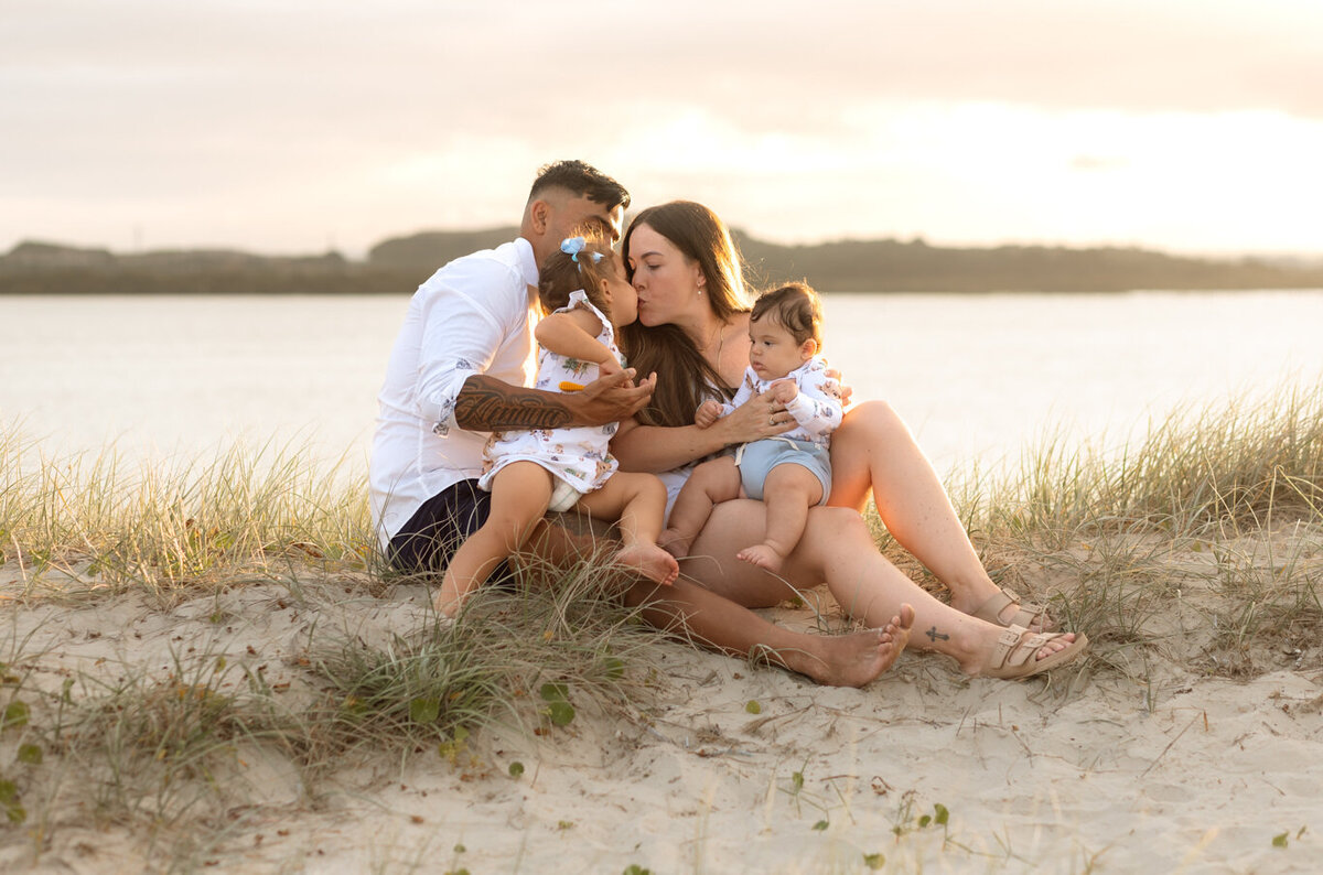 Family of four sitting on the sand by the water at sunset, with parents sharing a kiss while holding their young children in a tender, sunlit moment.