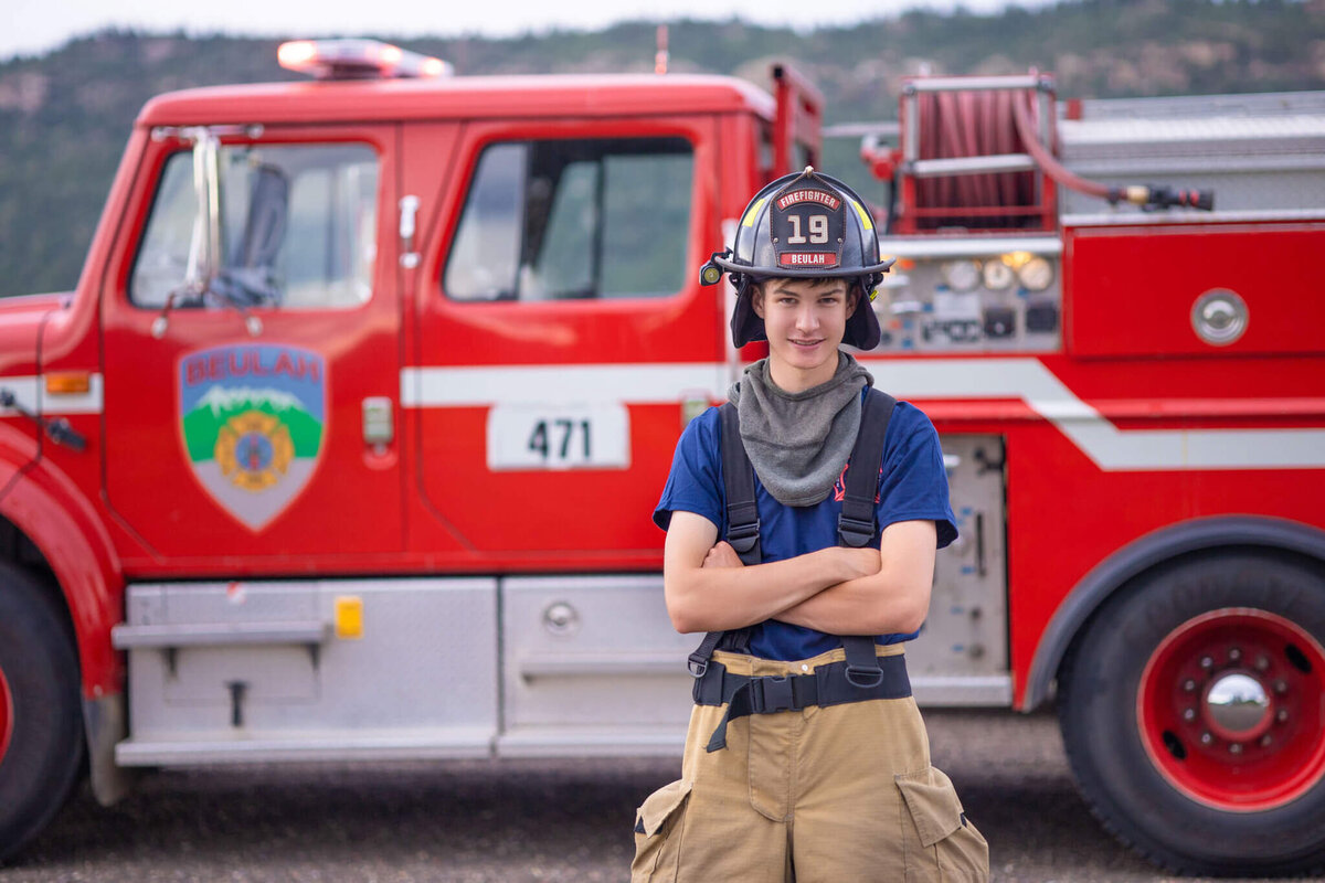 high school senior boy in bunker gear and firefigher hat standing in front of a firetruck