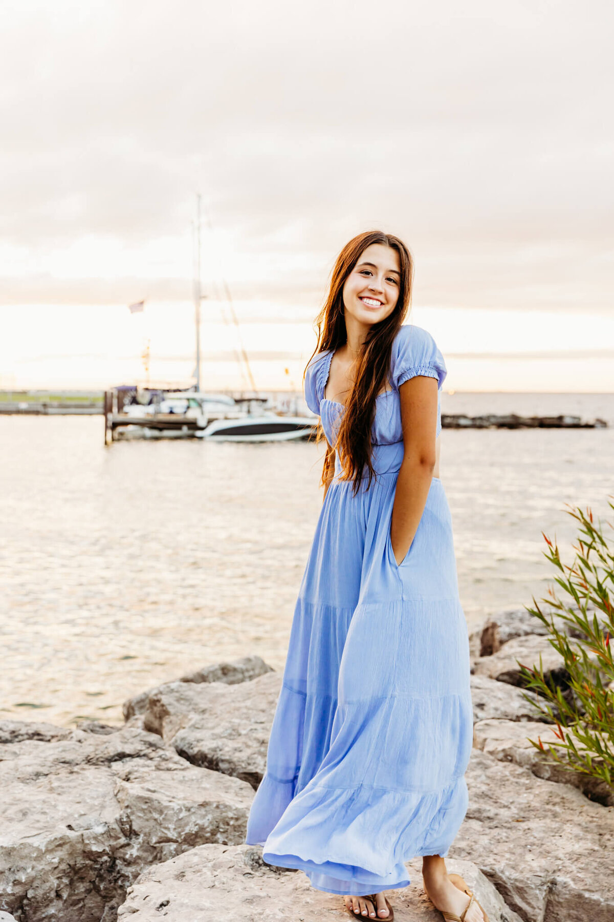 teenager twirling in her blue dress on top of large rocks with the lake and sailboats in the background
