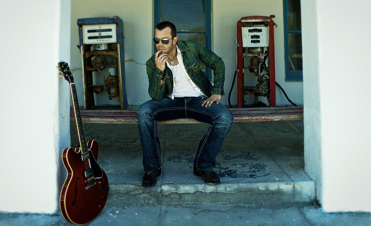Male musician portrait Aaron Pritchett wearing blue denim jacket white shirt blue jean pants with sunglasses while sitting between old gas pumps beside red electric guitar