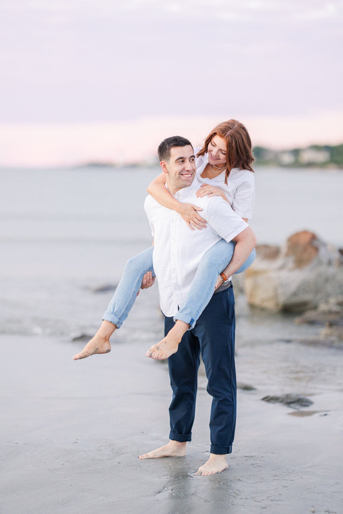 A man giving his fiance a piggyback ride on Wingaersheek Beach at sunset