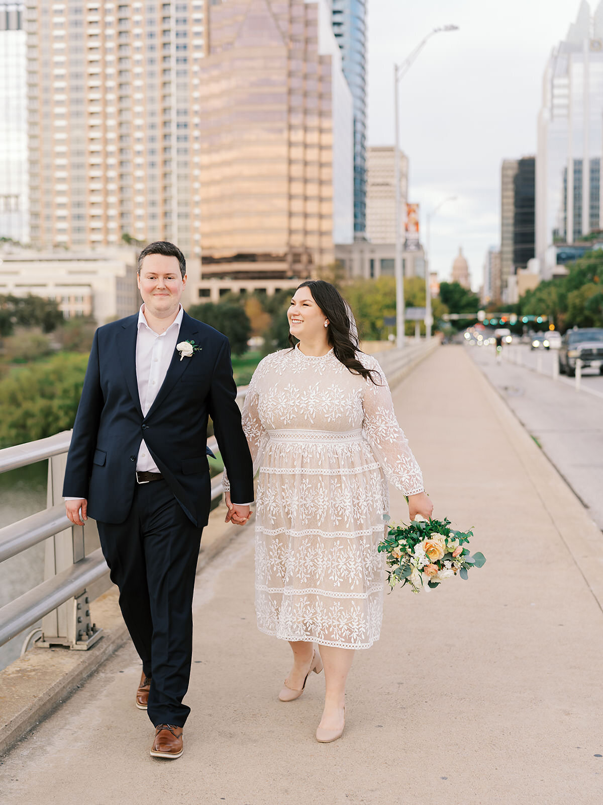 couple in white dress, navy suit, holding a bouquet walk across the South Congress bridge in Austin